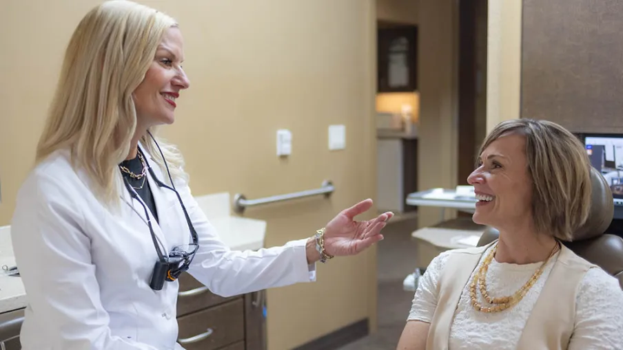 Female dentist in white coat consulting and smiling with a female patient in dental office chair.
