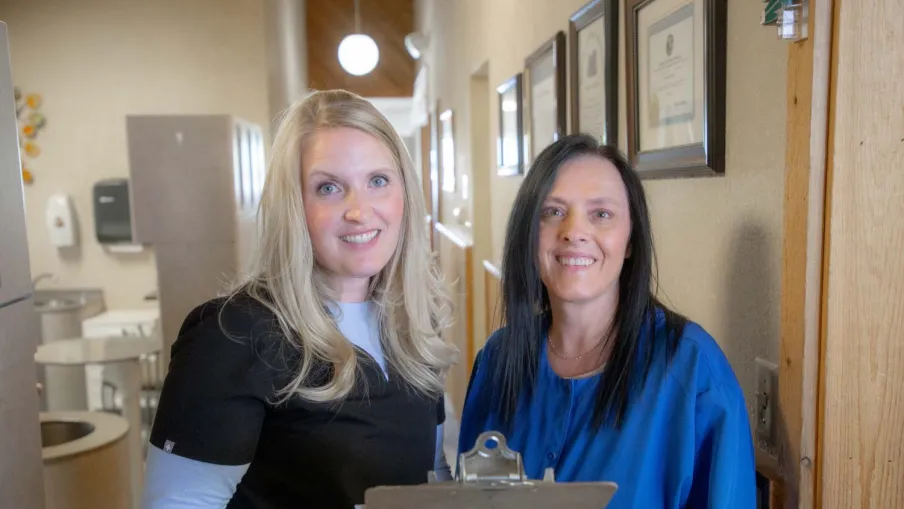 Two women in a medical office holding a clipboard and smiling, standing in a hallway with framed certificates.