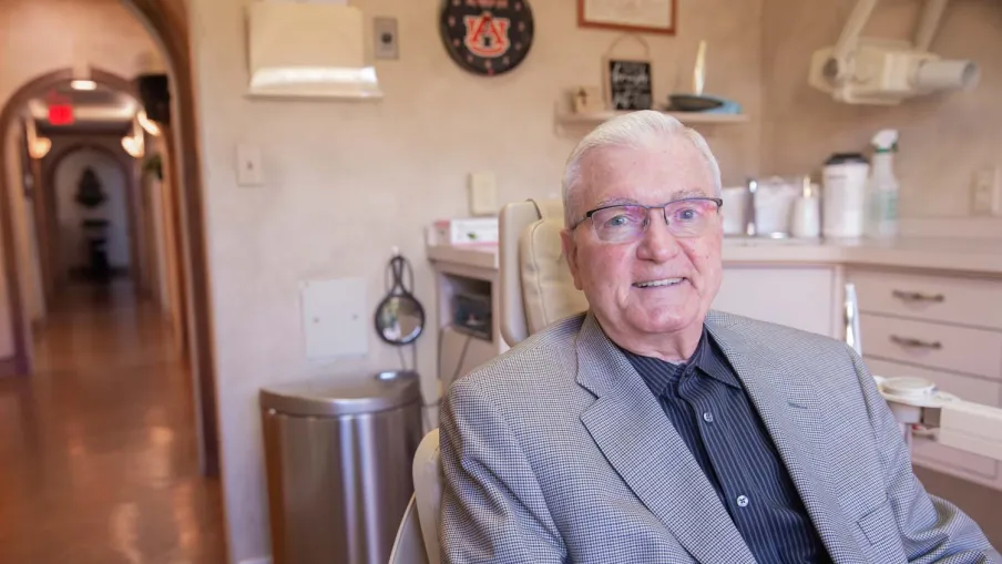 Elderly man in glasses and suit sitting in a well-lit medical or dental office with beige walls.