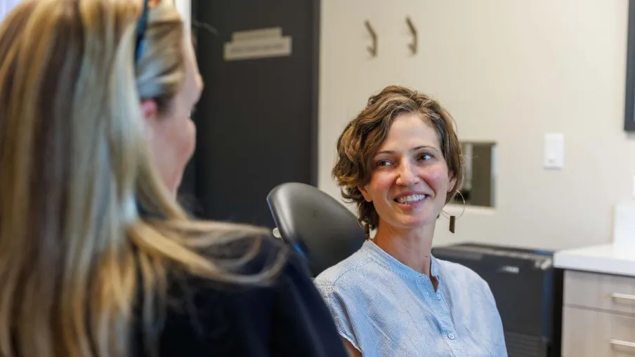 Two women in conversation in a modern office setting, one smiling and seated in a chair facing the other.