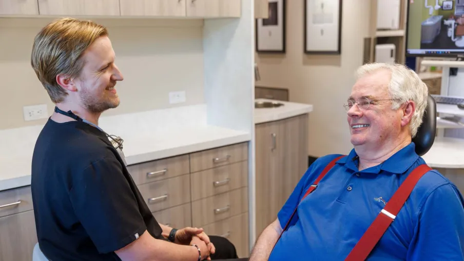 Dentist and elderly male patient smiling and talking in a modern dental office with wooden cabinets.