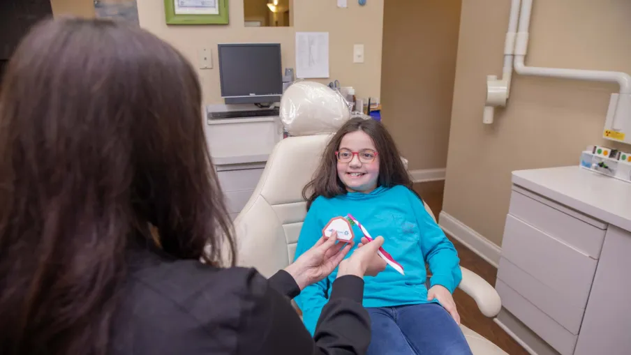 Young girl smiles in dental chair as dentist demonstrates proper brushing with a model and toothbrush