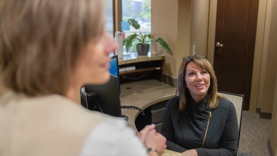 Smiling receptionist helping a visitor at the front desk in a modern office with plant and computer visible.