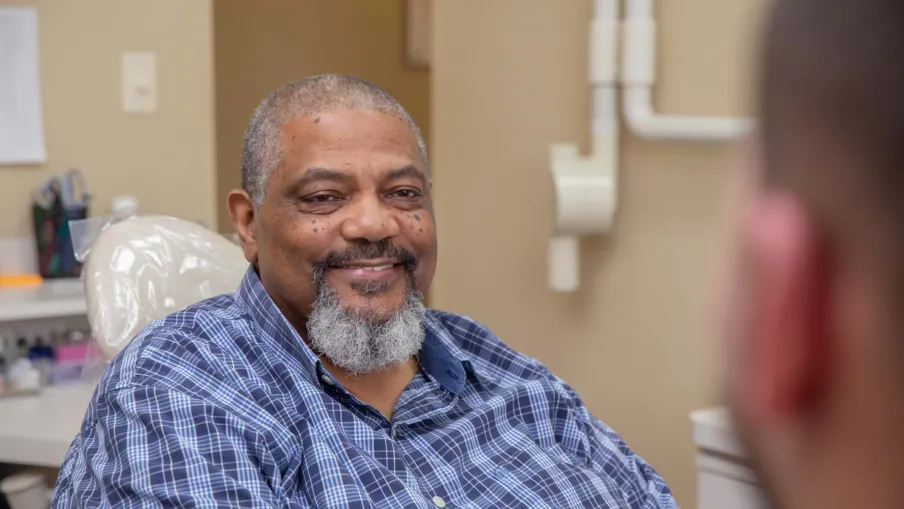 Smiling man with gray beard wearing a blue plaid shirt sitting and talking in a medical office.