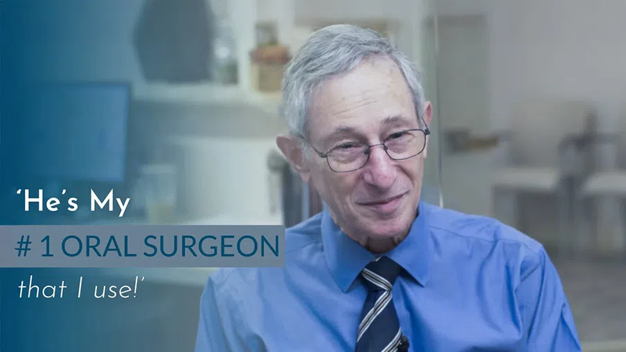 Smiling older man in glasses and blue shirt with text praising his oral surgeon in a dental office.