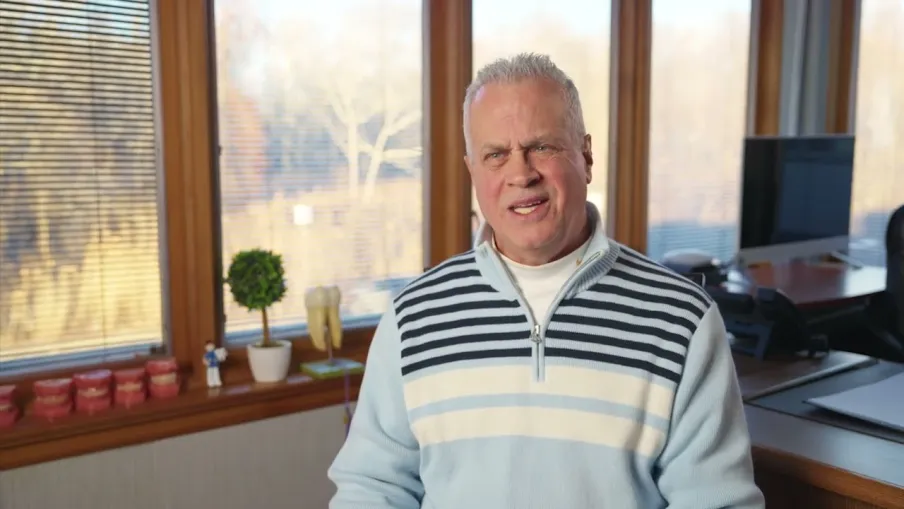 Man in a light blue striped sweater sitting in an office with large windows and wooden accents.