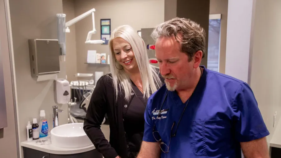 Dentist in blue scrubs consulting with female patient in modern dental clinic room with sink and equipment.