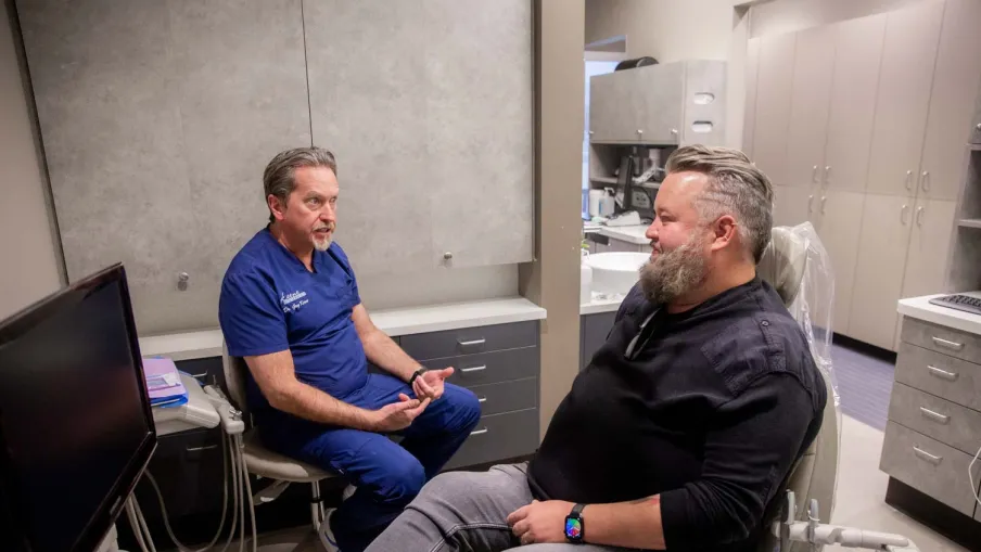 Dentist in blue scrubs consulting a male patient seated in a dental chair in a modern clinic room.