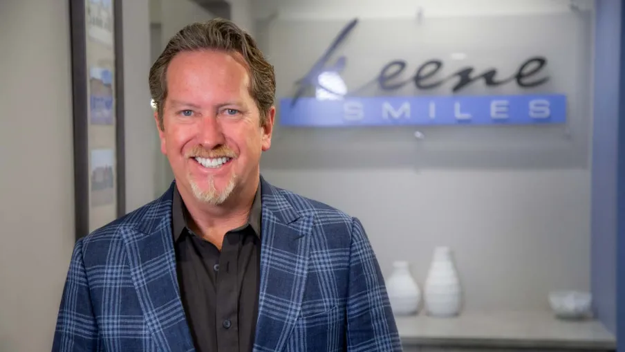 Smiling man in a blue plaid jacket standing inside a dental office with a Keene Smiles sign in the background