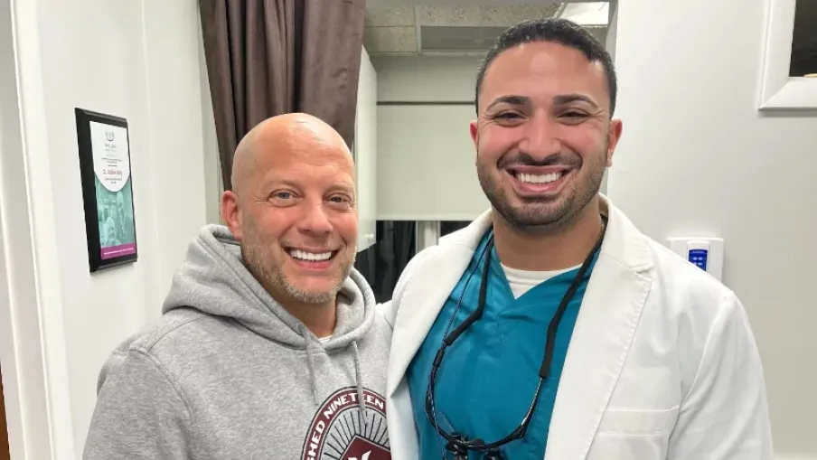 Smiling man in gray hoodie poses with a male dentist wearing white coat and teal scrubs in a clinic setting