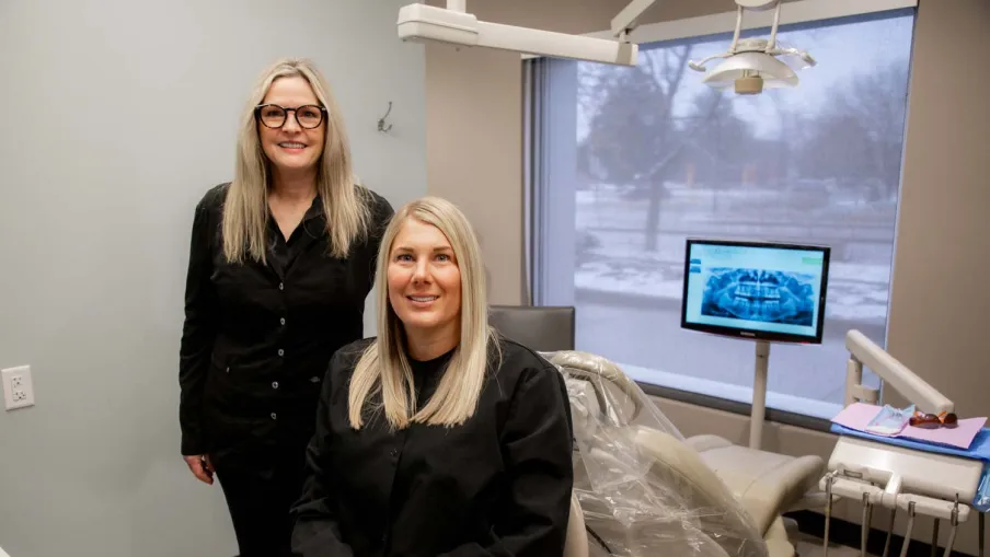 Two female dentists in black uniforms posing in a modern dental office with equipment and X-ray on screen.
