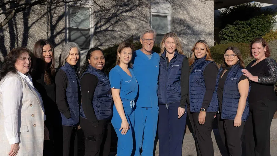 Group of medical and office staff posing outdoors in uniforms and casual wear in front of a building in sunlight.