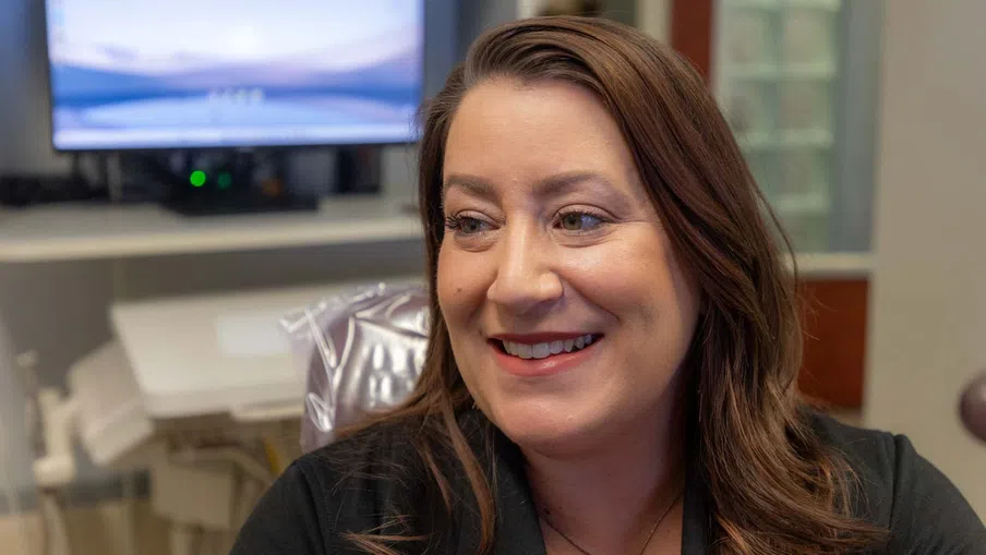 Smiling woman with brown hair wearing black shirt in a modern office setting with computer screen in background