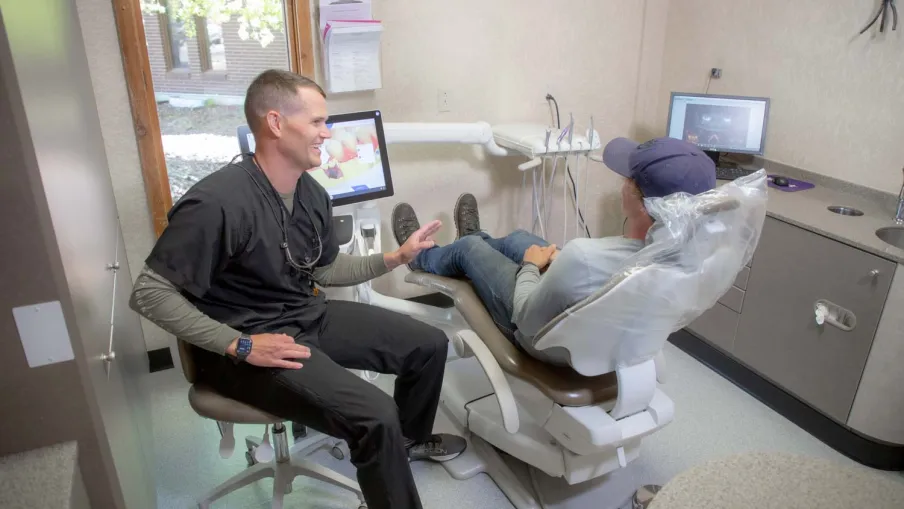 Dentist in black scrubs talking to a patient reclined in a dental chair during a consultation.