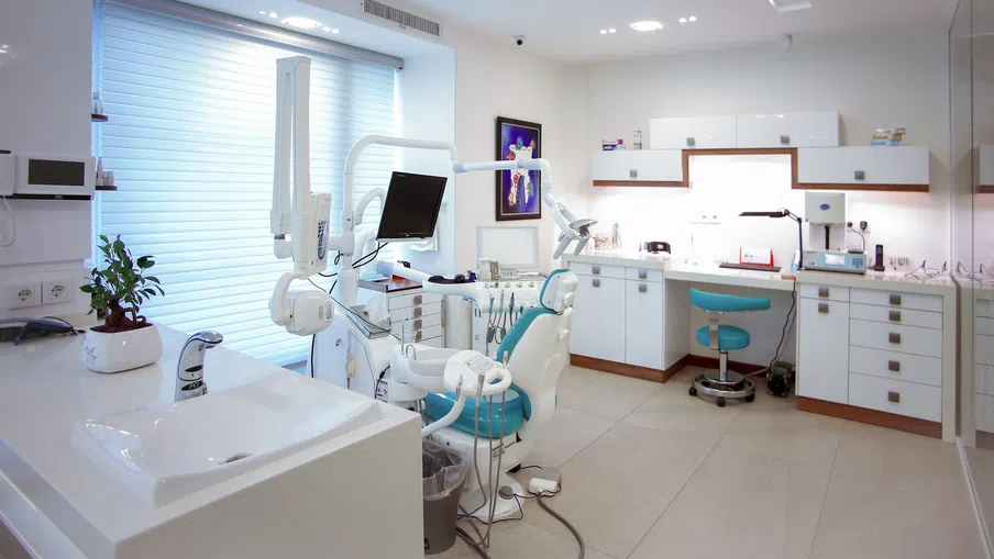 Bright modern dental clinic with dental chair, monitor, sink, and white cabinetry under natural light.