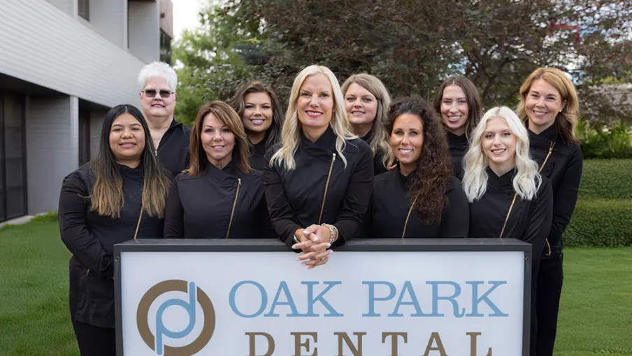 Dental team of nine women standing behind Oak Park Dental sign outside building with greenery.