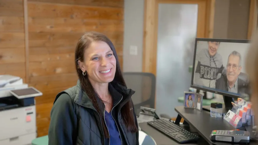 Smiling woman with long hair in office setting with computer, keyboard, mouse, and wooden wall background.