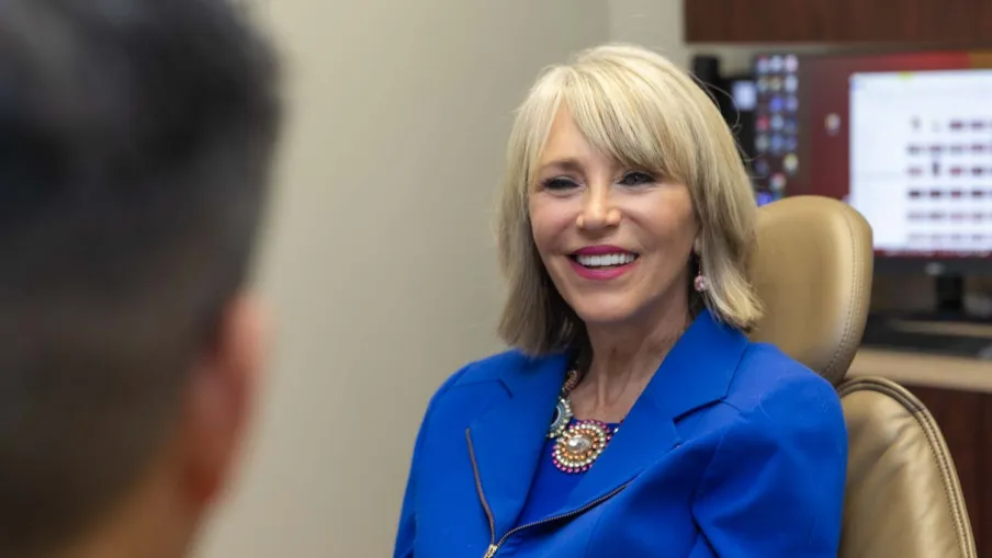 Smiling woman in a blue blazer sitting in an office chair during a meeting with a blurred person in the foreground
