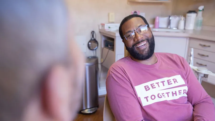 Smiling man in glasses wearing a pink sweatshirt that says Better Together talking indoors with another person.