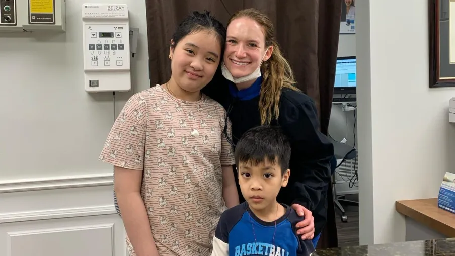 Smiling woman with two children in a medical office holding a plush toy, standing near a curtain and countertop.