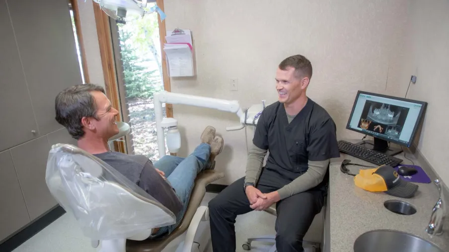 Dentist in black scrubs consulting a male patient seated in a dental chair in a bright clinic room.