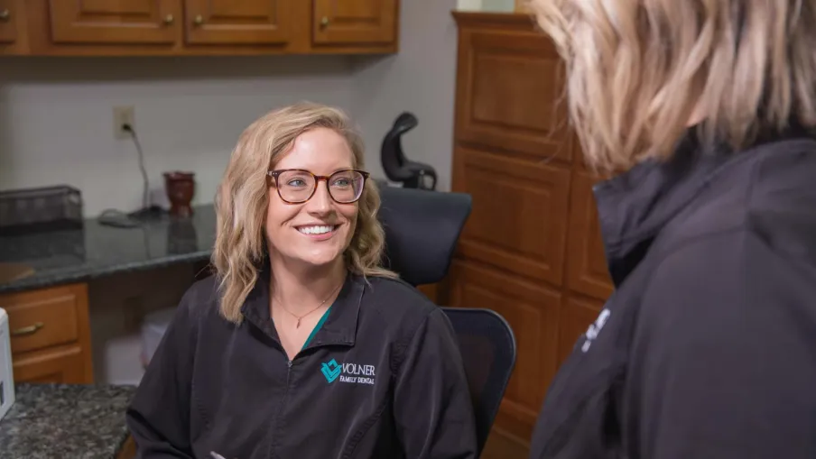 Smiling female dental professional in a clinic wearing Volner Family Dental uniform and glasses talking to colleague.