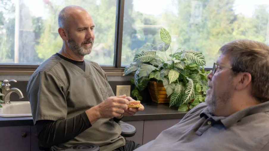 Dentist explains dental implant model to patient in clinic with plant and window background
