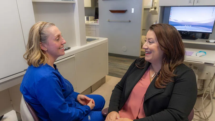 Two women smiling and talking in a medical office, one wearing blue scrubs and the other in business attire.
