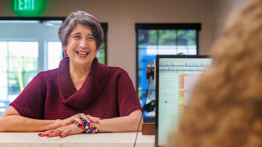 Smiling woman in burgundy sweater sitting at a reception desk with computer screen and window in background