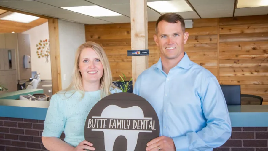 Man and woman holding a Butte Family Dental sign inside a dental office with wood panel walls.