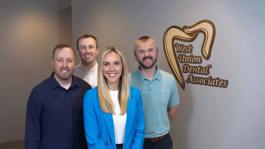 Four dental professionals smiling inside West Union Dental Associates office with logo on wall.