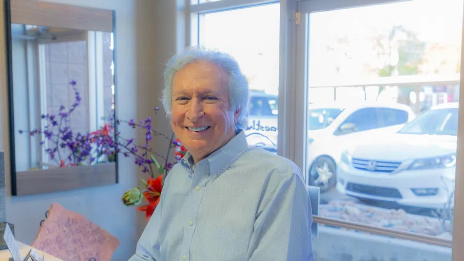 Smiling elderly man in light blue shirt sitting indoors near window with flowers and parked cars outside.