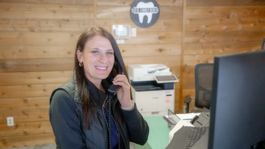 Smiling woman with dark hair on phone at desk in office with wooden walls and computer monitor