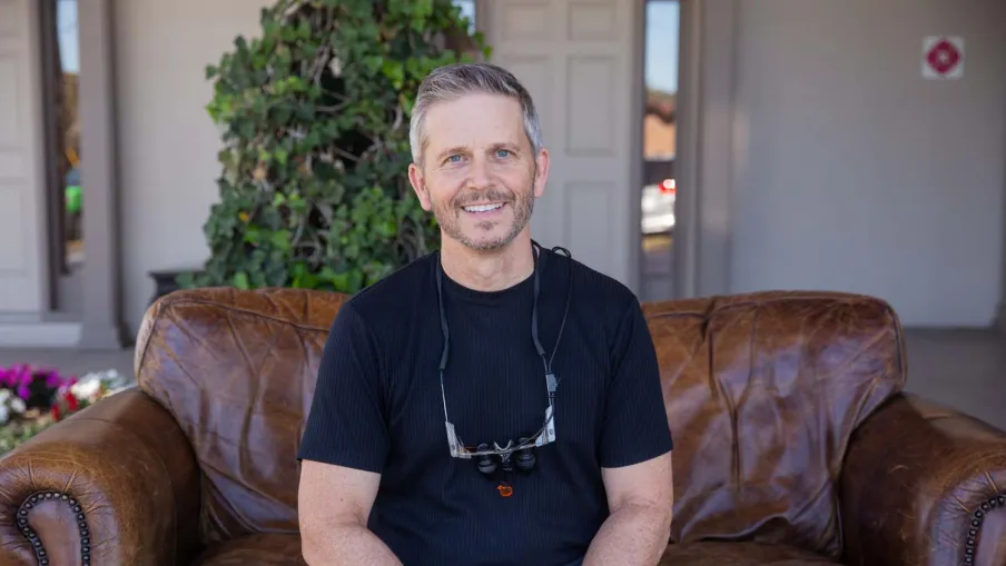 Smiling man in black shirt with dental loupes around neck sitting on brown leather couch outdoors.