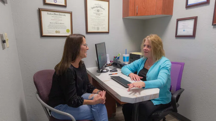 Two women having a professional conversation in a small office with diplomas on the wall and computer on desk