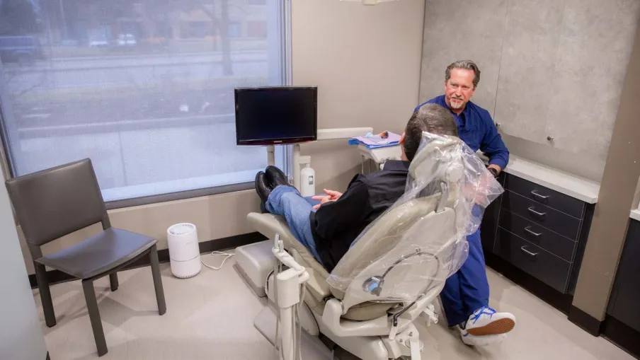 Dentist talking to patient sitting in dental chair in a modern dental office room with large window.