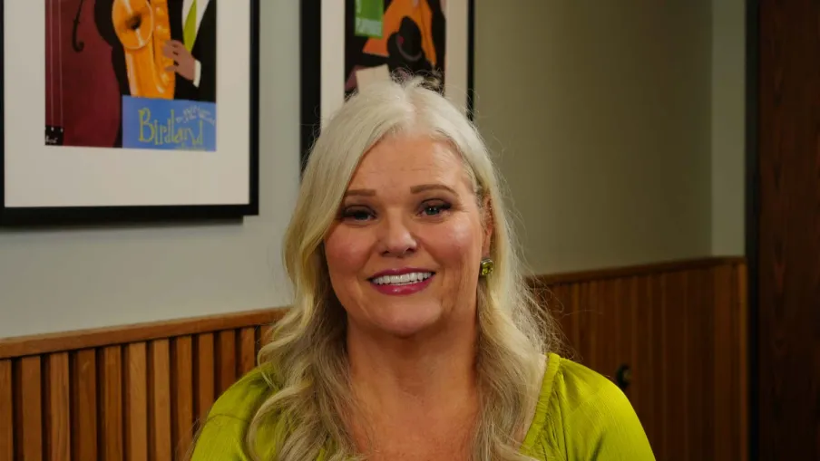 Smiling woman with long blonde hair wearing a green top seated indoors with framed art behind her.