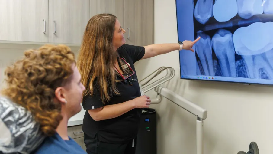 Dentist explaining dental X-ray results to patient showing teeth and roots on a monitor in clinic.