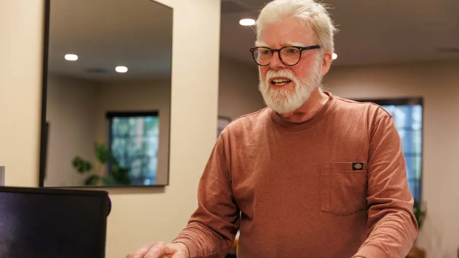 Elderly man with glasses smiling while using a laptop in a cozy modern home interior.