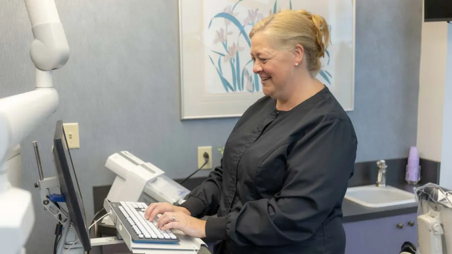 Smiling dental professional in black scrubs typing on a keyboard in a modern dental office setting