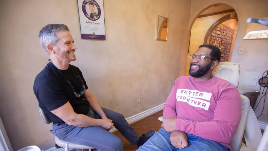 Two men smiling and talking comfortably in a cozy dental office setting with dental equipment visible.
