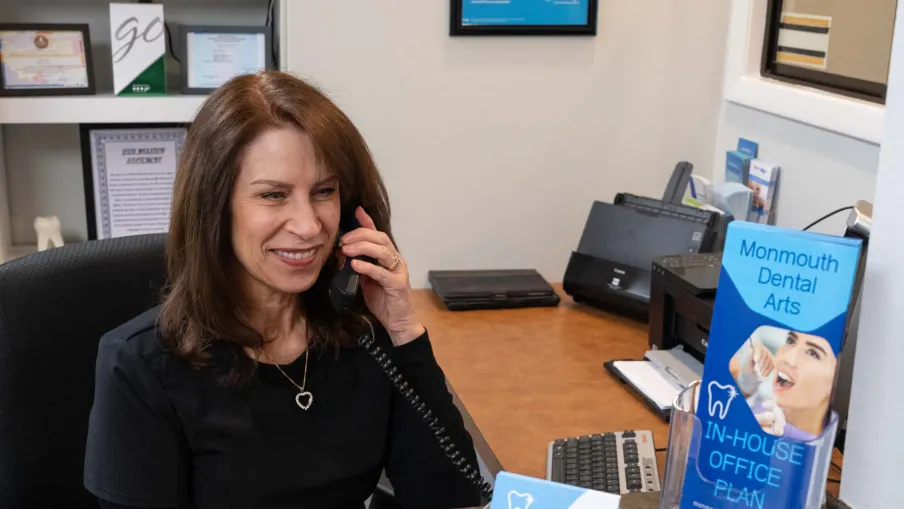 Receptionist smiling while speaking on the phone at a dental office with brochures displayed on the counter.