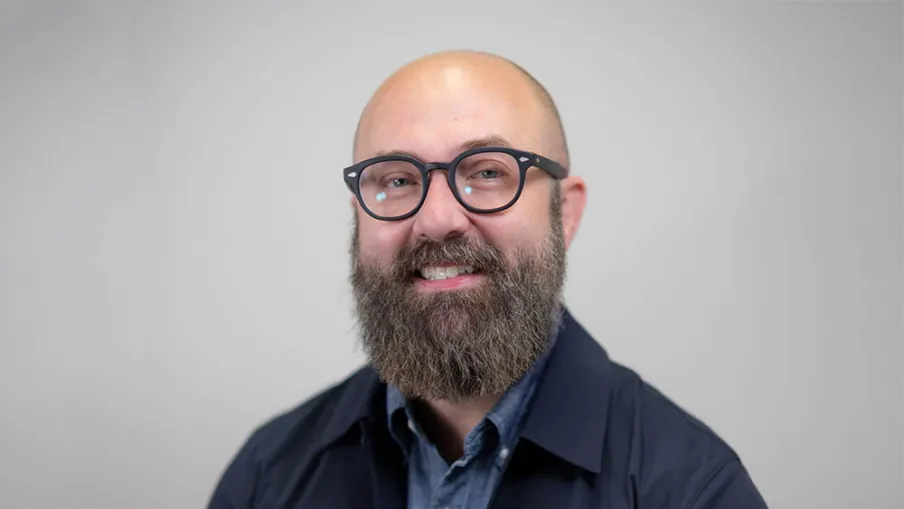 Smiling bald man with a full beard wearing glasses, a blue shirt, and a dark jacket against a plain background