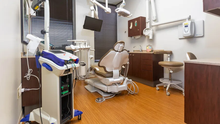 Modern dental office with beige dental chair, medical equipment, wooden flooring, and dark window blinds.
