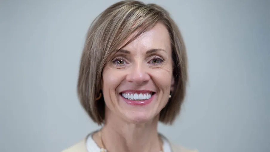 Smiling woman with short blonde hair wearing a white top and beige vest against a gray background