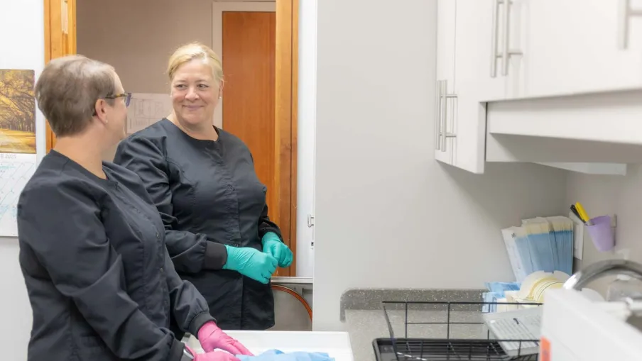 Two healthcare workers in black scrubs and gloves organizing medical supplies in a clinic room with white cabinets.