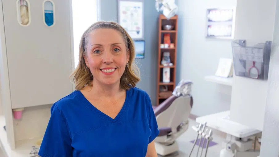 Smiling female dentist in blue scrubs standing in a modern dental clinic with dental chair and tools.