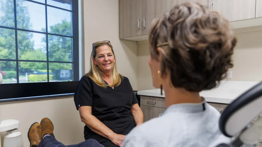 Dental hygienist converses with patient seated in dental chair in a bright clinic room with large window.
