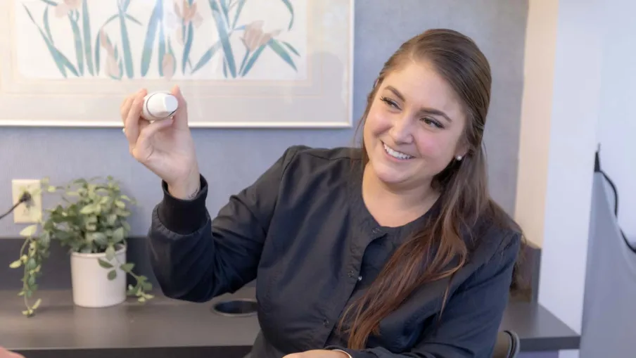 Smiling dental hygienist in black jacket showing a dental product to a patient in a clinic room.