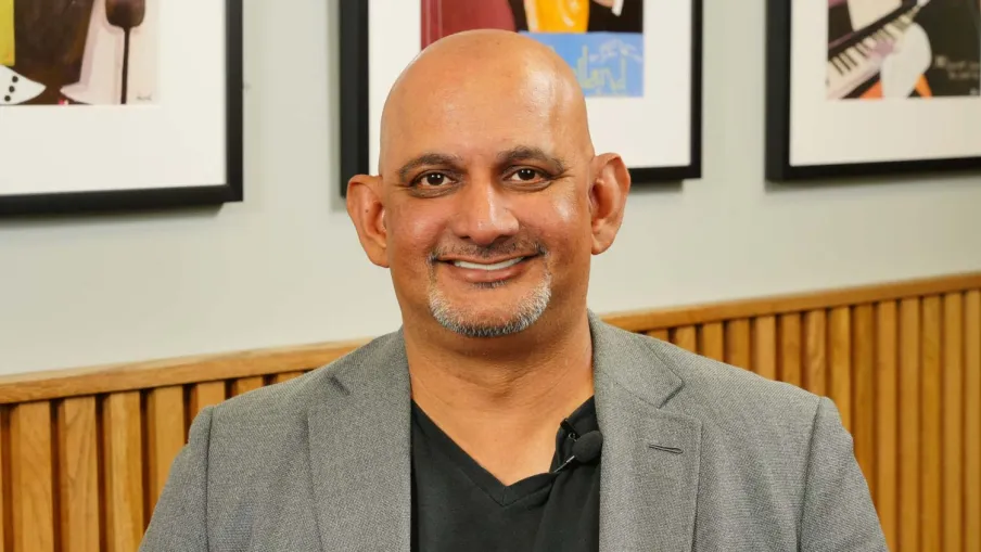 Smiling bald man with a goatee wearing a gray blazer and black shirt sitting indoors with framed art behind.